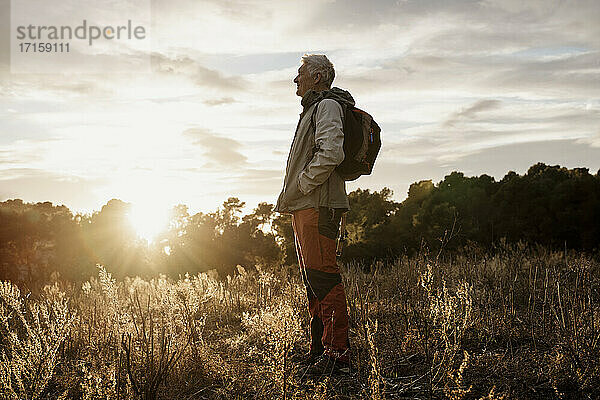 Älterer männlicher Wanderer mit Händen in den Taschen genießt die Aussicht auf den Sonnenuntergang auf einem landwirtschaftlichen Feld