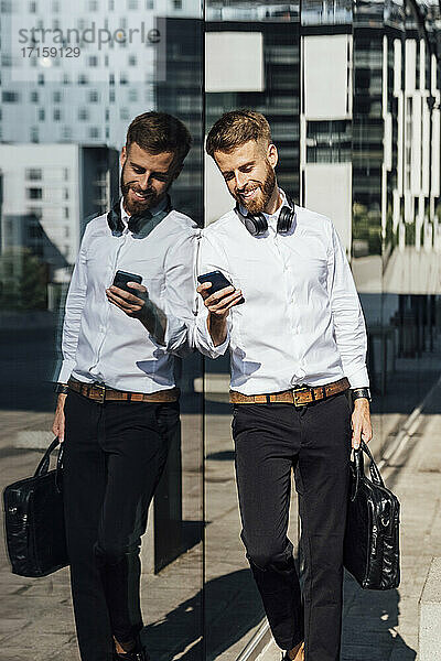 Smiling businessman with briefcase using smart phone while leaning on glass wall