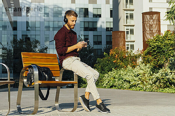 Unternehmer mit Kopfhörern  der ein Mobiltelefon benutzt  während er auf einer Bank in der Stadt sitzt
