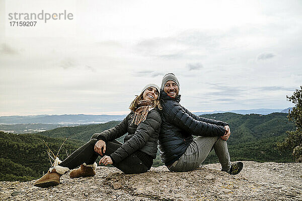 Lächelndes Paar sitzt Rücken an Rücken auf dem Aussichtspunkt gegen den Himmel