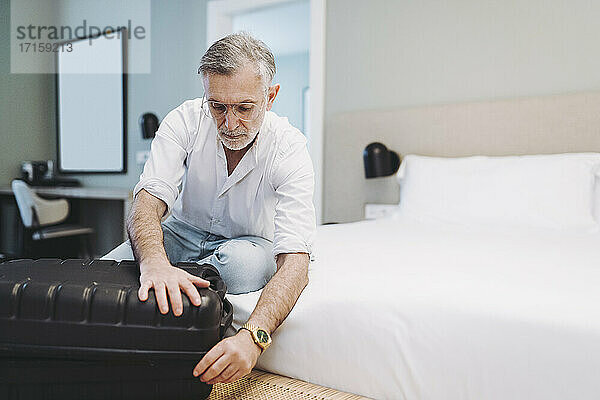 Man closing suitcase while sitting on bed in hotel room