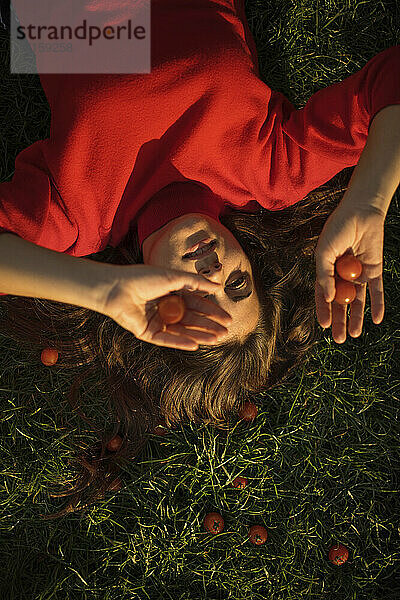 Junge Frau in rotem Kleid hält Kirschtomaten im Gras liegend an einem sonnigen Tag