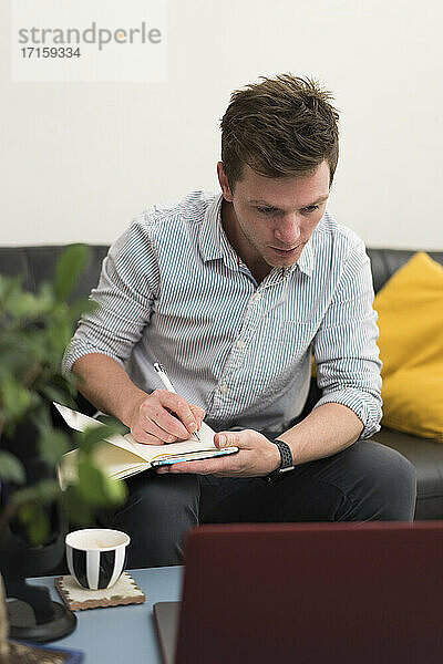 Geschäftsmann schreibt in ein Buch  während er im Büro zu Hause mit seinem Laptop arbeitet