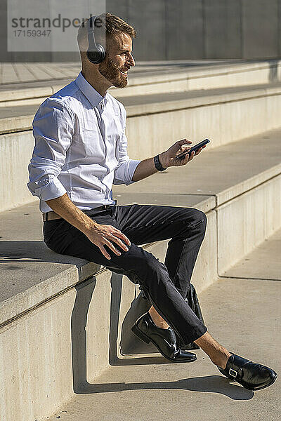 Smiling businessman listening music through headphones while using mobile phone sitting on steps