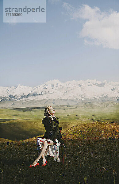 Beautiful woman sitting on chair while looking away against cloudy sky