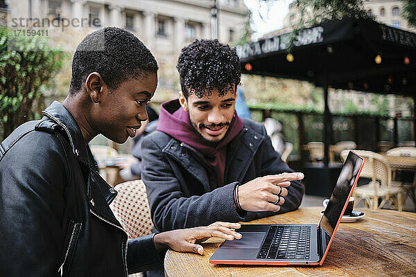 Man pointing toward laptop while sitting by friend at sidewalk cafe