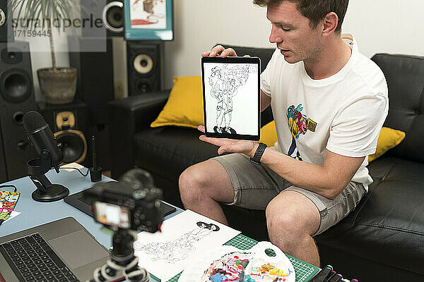 Männlicher Künstler zeigt digitales Tablet  während er zu Hause auf seinem Laptop live streamt