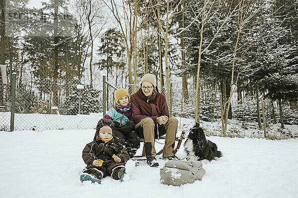 Mutter mit Töchtern und Border Collie auf verschneitem Feld vor Bäumen sitzend