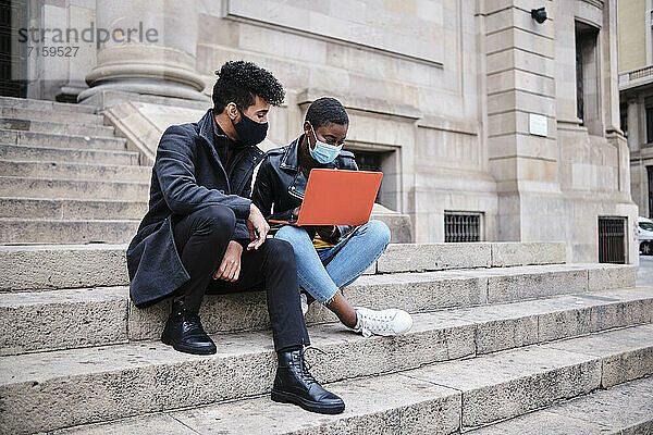 Friends wearing protective face mask using laptop while sitting on steps in city