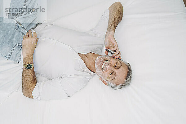 Smiling man talking on smart phone while lying on bed