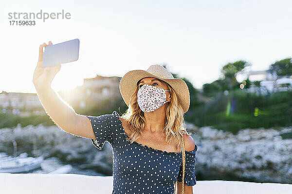 Ältere Frau mit Gesichtsmaske macht ein Selfie mit ihrem Smartphone im Dorf Binibeca  Menorca  Spanien