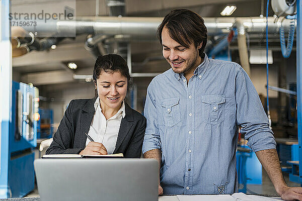 Smiling businesswoman working over laptop with colleague while standing at industry