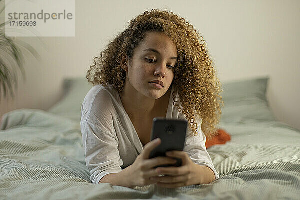 Junge Frau  die im Schlafzimmer auf dem Bett liegend eine Textnachricht über ihr Mobiltelefon sendet