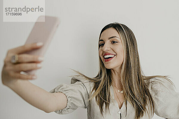 Smiling beautiful woman taking selfie through smart phone against white background