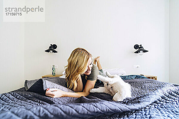 Blonde Frau spielt mit Welpe auf dem Bett gegen weiße Wand zu Hause