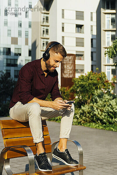Geschäftsmann mit Kopfhörern  der sein Smartphone benutzt  während er auf einer Bank sitzt