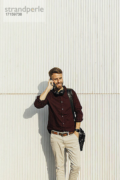Lächelnder Geschäftsmann mit Umhängetasche  der mit seinem Handy telefoniert  während er an einer weißen Wand steht