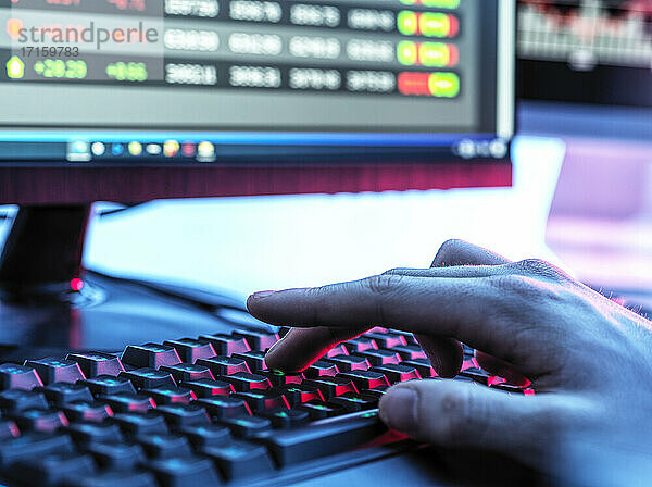 Hände eines Händlers  der auf einer Tastatur vor einem Computermonitor tippt  auf dem Börsendaten angezeigt werden