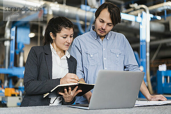 Young businesswoman writing in book while working with colleague at industry
