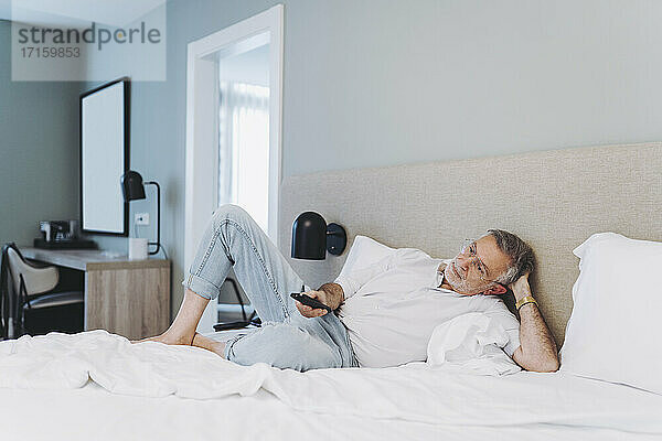Relaxed man using remote control while lying on bed in hotel room