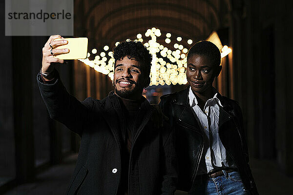 Smiling man taking selfie through mobile phone while standing with friend at corridor
