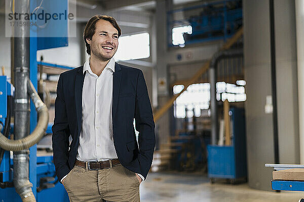 Smiling businessman looking away while standing with hands in pockets at industry