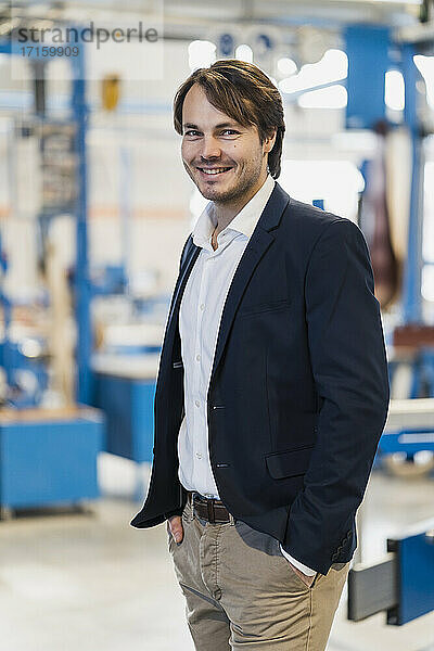 Male entrepreneur smiling while standing with hands in pockets at industry