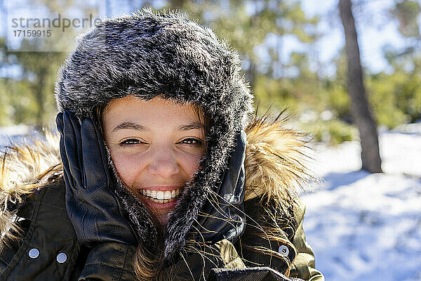 Fröhliche Frau mit Pelzmütze  die im Winter lächelnd im Wald steht