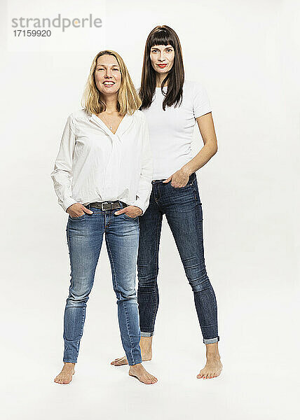 Female friends with hands in pockets standing against white background