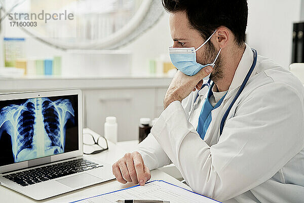 Thoughtful male doctor analyzing X-ray image on laptop while sitting in clinic