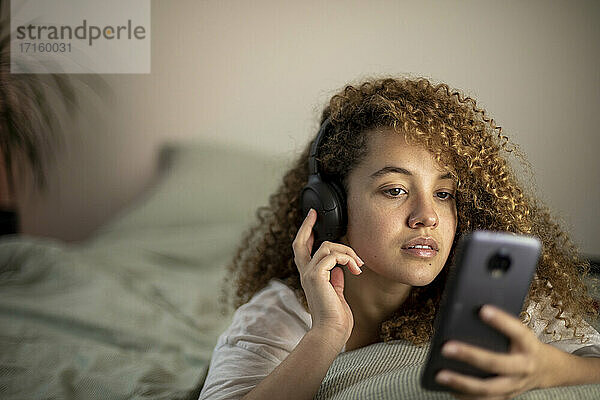 Junge Frau auf dem Bett liegend  während sie ein Smartphone benutzt und zu Hause Musik hört