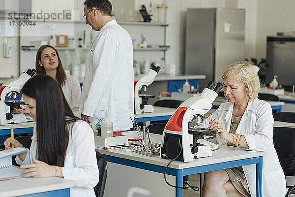 Ältere Forscherin im weißen Kittel bei der Arbeit im Labor  umgeben von anderen Forschern