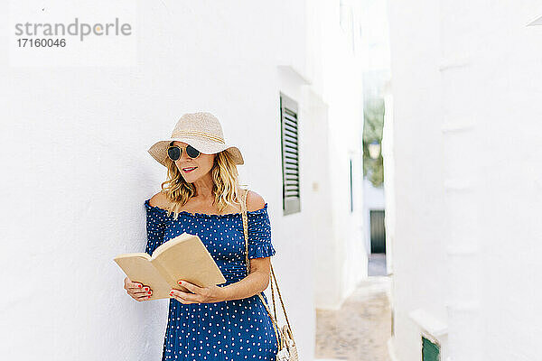 Frau mit Hut liest ein Buch  während sie bei einem Haus im Dorf Binibeca  Menorca  Spanien  steht
