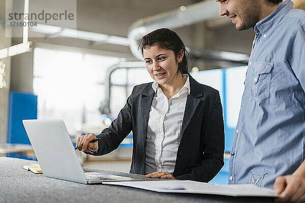 Young businesswoman working on laptop while standing with colleague at industry