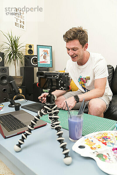 Content-Creator zeichnet auf Skizzenblock  während er mit seinem Laptop bei einem Live-Streaming zu Hause sitzt
