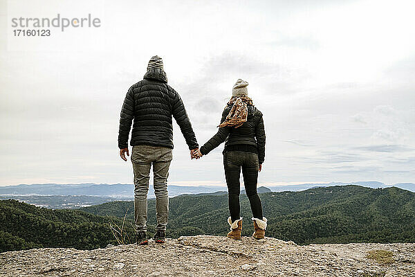 Paar  das sich an den Händen hält  während es an einem Aussichtspunkt steht und die Berge gegen den Himmel betrachtet