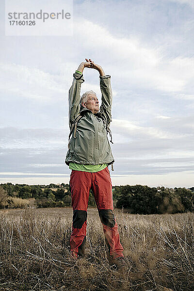 Älterer männlicher Wanderer  der die Arme ausstreckt  während er auf einem landwirtschaftlichen Feld vor einem bewölkten Himmel steht