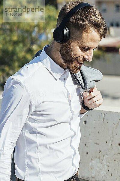 Smiling businessman wearing headphones looking down while standing outdoors