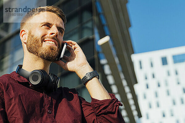 Lächelnder Geschäftsmann mit Kopfhörern  der in der Stadt mit seinem Mobiltelefon spricht