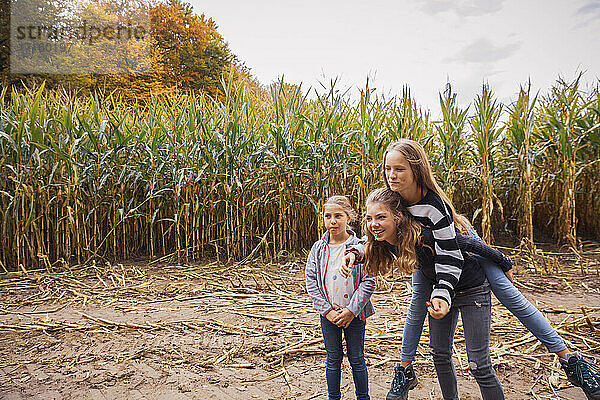 Smiling girl piggybacking friends while standing at corn field