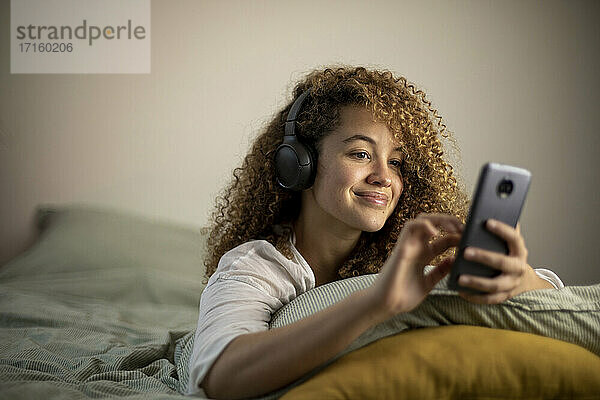 Lächelnde junge Frau  die Musik hört und ein Mobiltelefon benutzt  während sie zu Hause auf dem Bett liegt