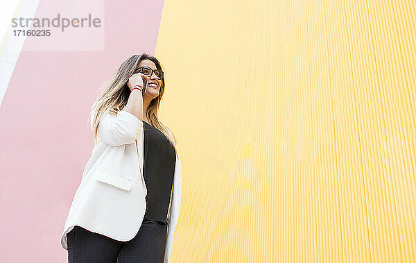 Smiling young woman talking on phone against pink and yellow wall