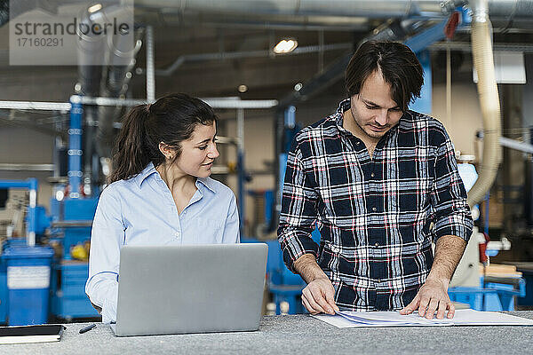 Businesswoman with laptop discussing over document while standing by colleague at industry