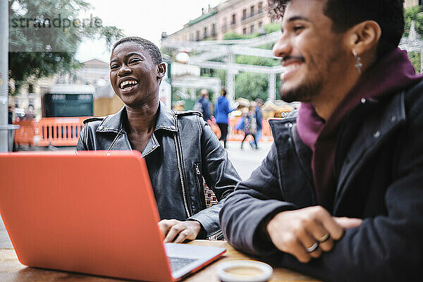 Young woman with laptop smiling while sitting by friend at sidewalk cafe
