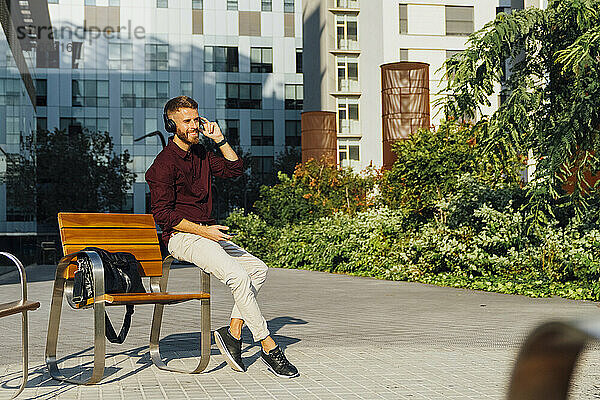 Lächelnder Geschäftsmann mit Mobiltelefon  der Kopfhörer einstellt  während er auf einer Bank in der Stadt sitzt