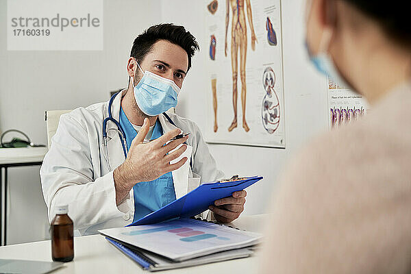 Male doctor with protective face mask talking with patient in clinic