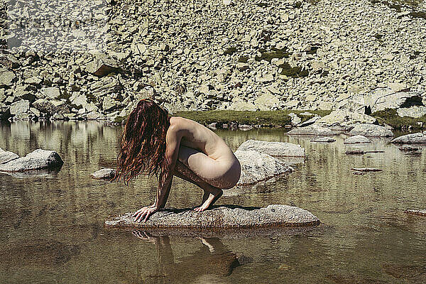 Young naked woman crouching on rock in lake during sunny day