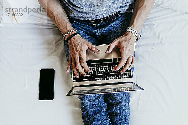 Draufsicht auf einen jungen Mann  der zu Hause auf dem Bett sitzend an einem Laptop und einem Mobiltelefon arbeitet