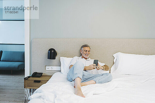 Happy man using smart phone while lying on bed against headboard in hotel room