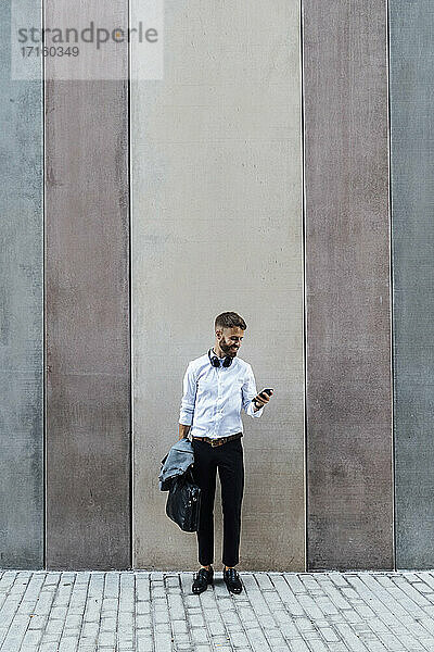 Businessman with briefcase and jacket using mobile phone while standing against wall